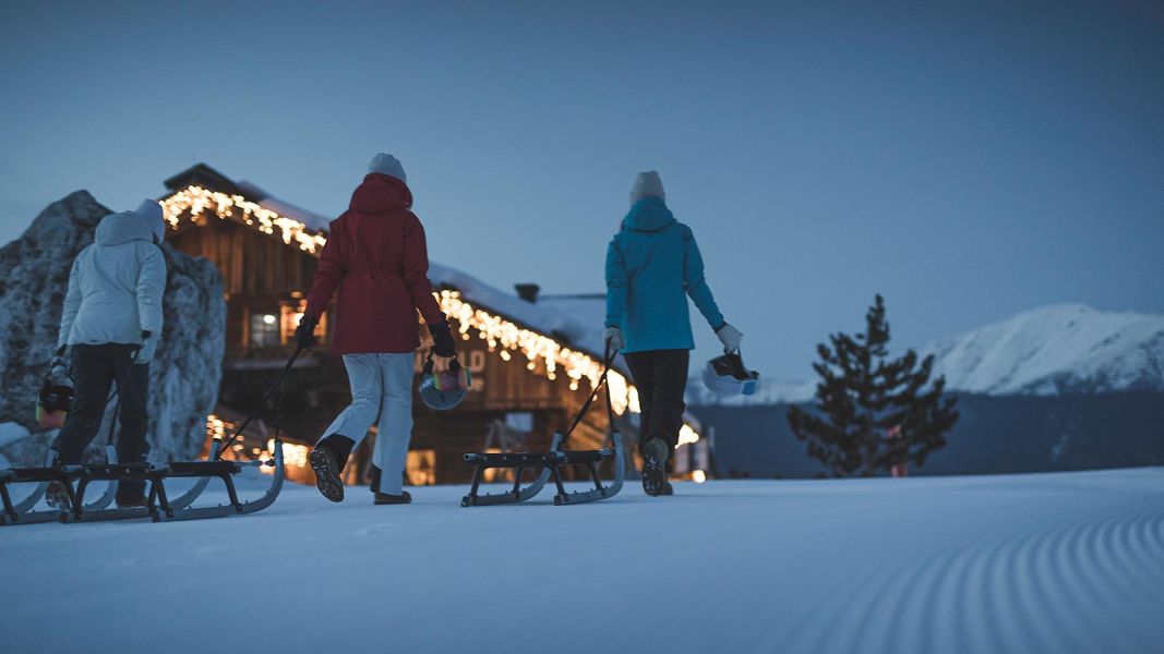 Tre persone che camminano nella neve trainando le slitte verso una baita illuminata al tramonto