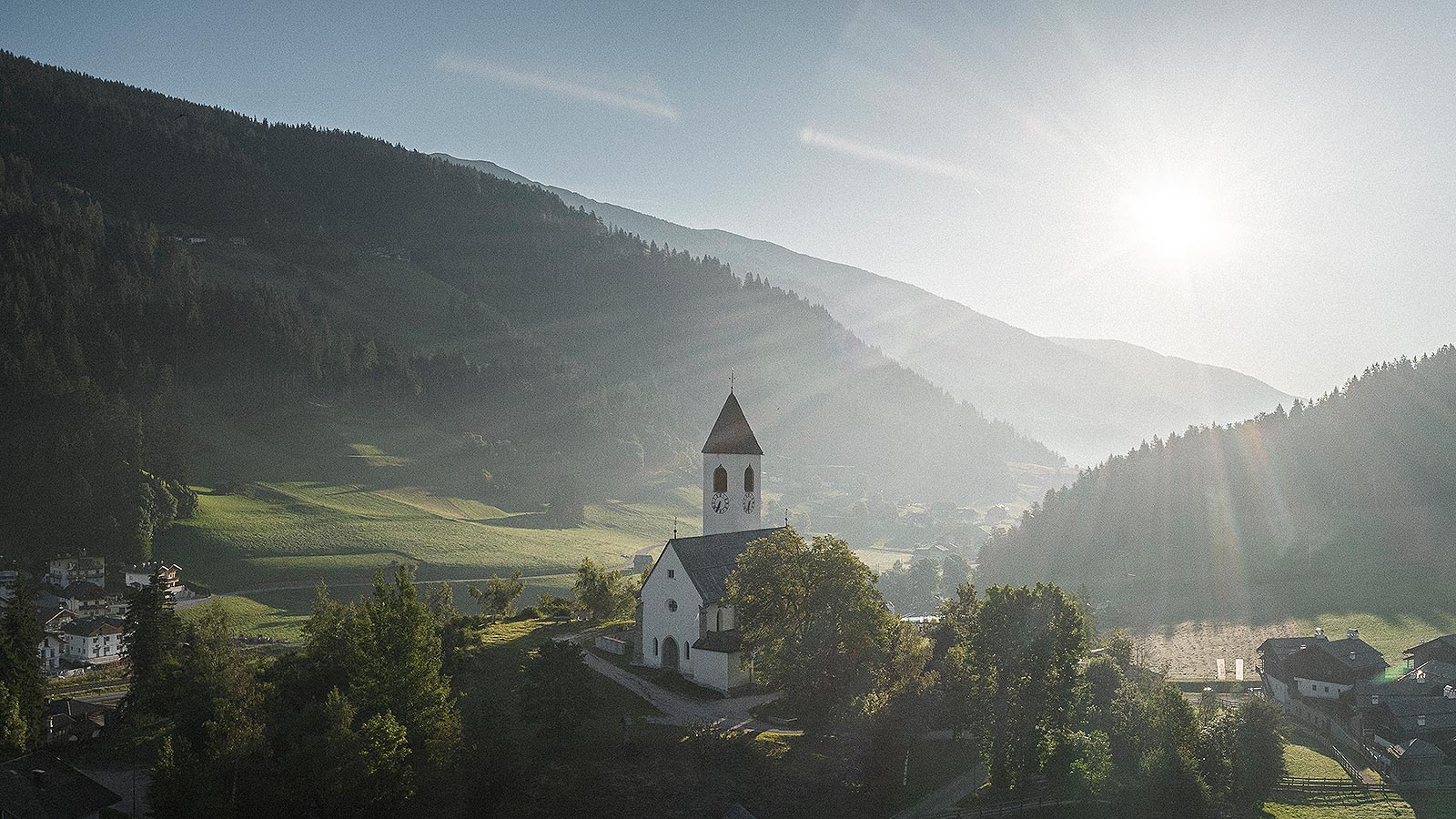 Kleine weiße Kirche mit Glockenturm inmitten der Pustertaler Hügellandschaft, die am späten Nachmittag von der Sonne beleuchtet wird.