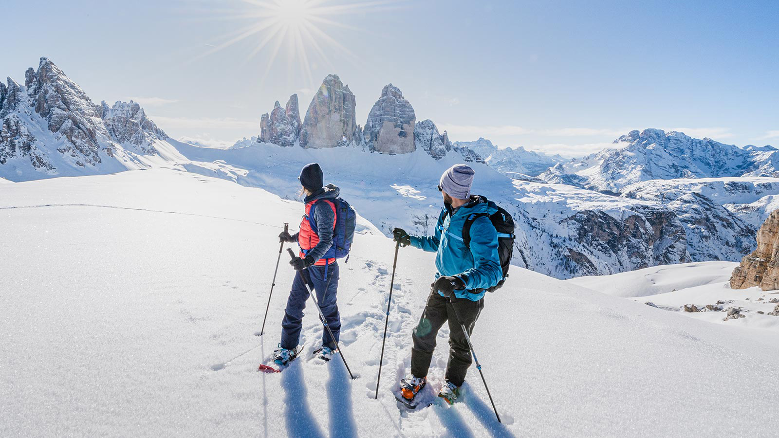 Ein paar Skifahrer gehen mit ihrer Skiausrüstung über die verschneiten Wege, während die Sonne auf den Berg scheint.