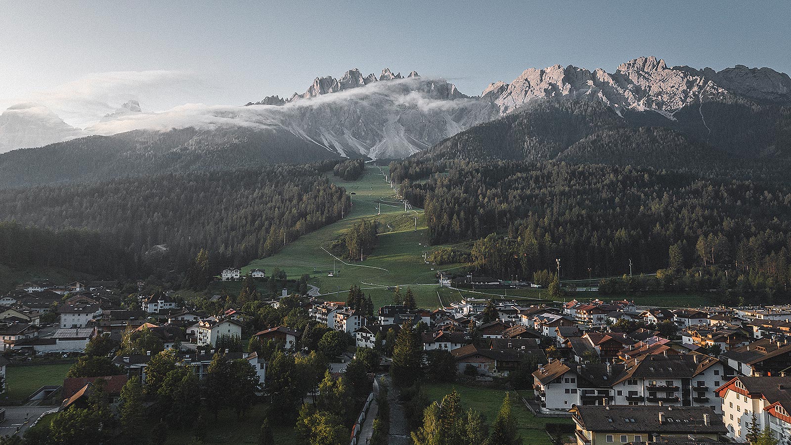 Panoramablick auf das Dorf Innichen im Vordergrund und die schneebedeckten Berge im Hintergrund, die mit der Standseilbahn in der Bildmitte erreicht werden können.